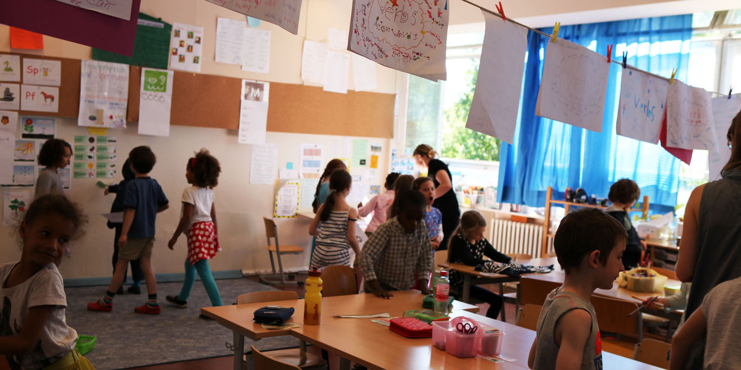 Ein Klassenraum mit mehreren Schülern und einer Lehrerin, in dem Raum hängt eine Wäscheleine mit mehreren Plakaten