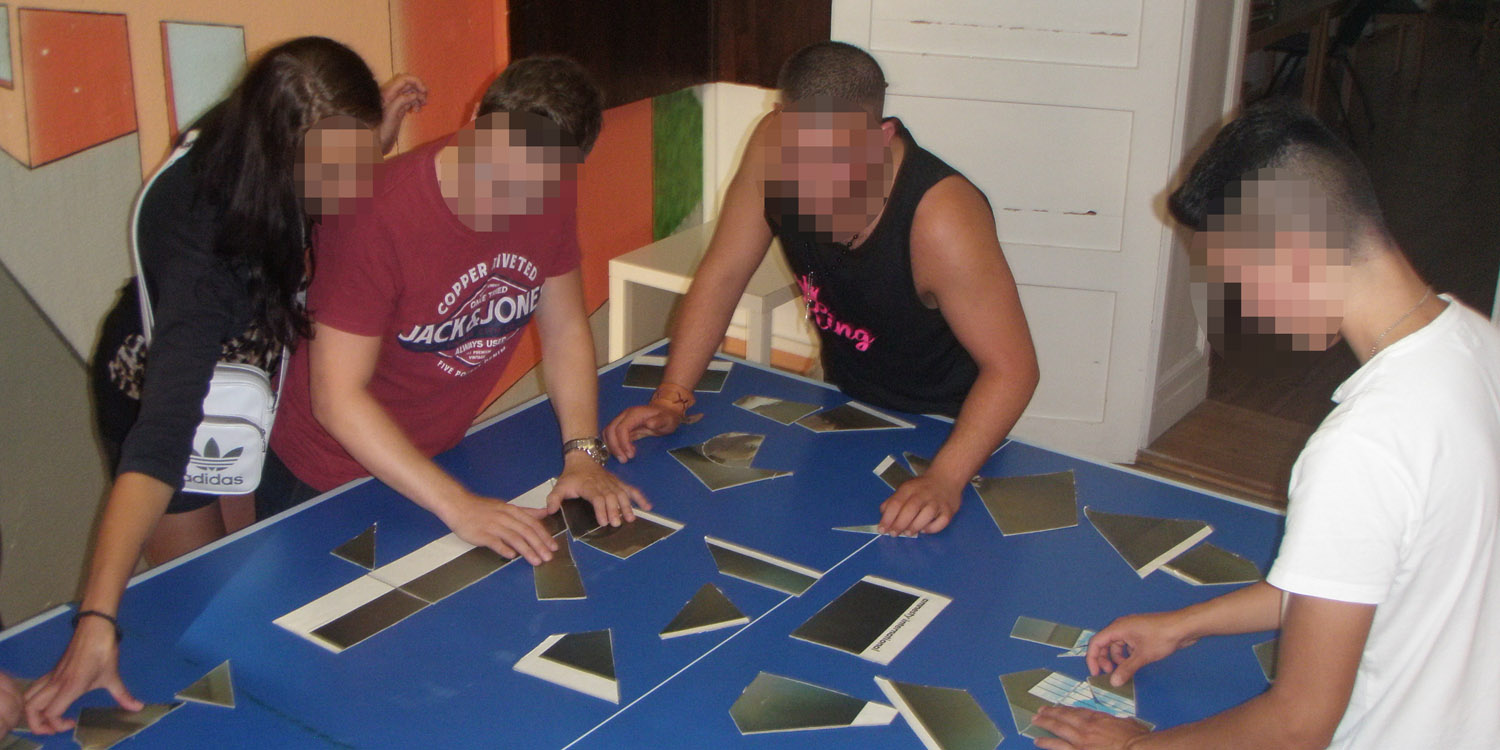 Zwei junge Männer stehen an einem Tisch und puzzeln aus verschiedenen Ausschnitten ein Bild zusammen.
