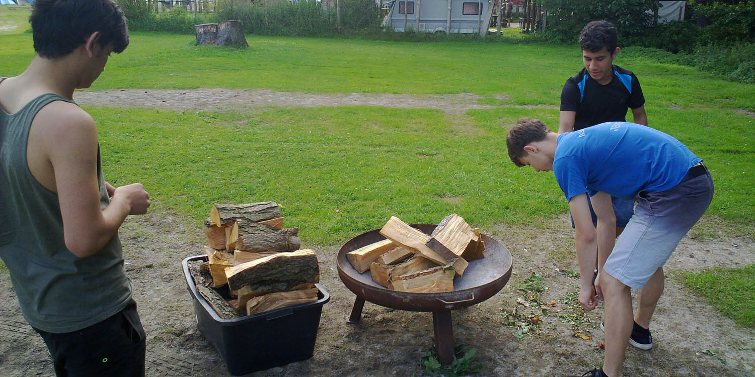 Drei Personen stehen um eine Metallschale, in der Schale sind mehrere Holzscheiten und daneben steht auch noch eine Kiste mit Holzscheiten