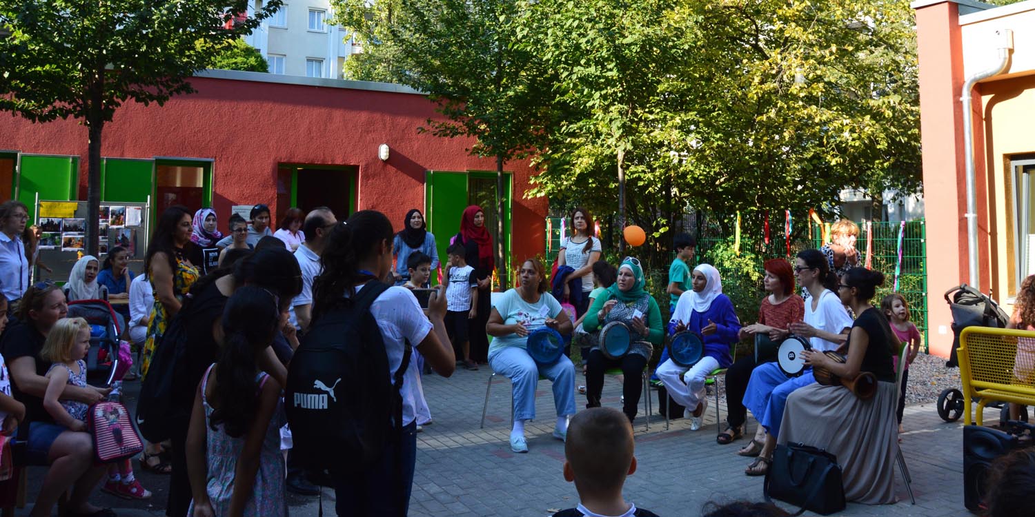 In der Mitte spielt eine Band mit einer Flöte und mehreren Trommeln und ringsherum stehen Erwachsene und Kinder