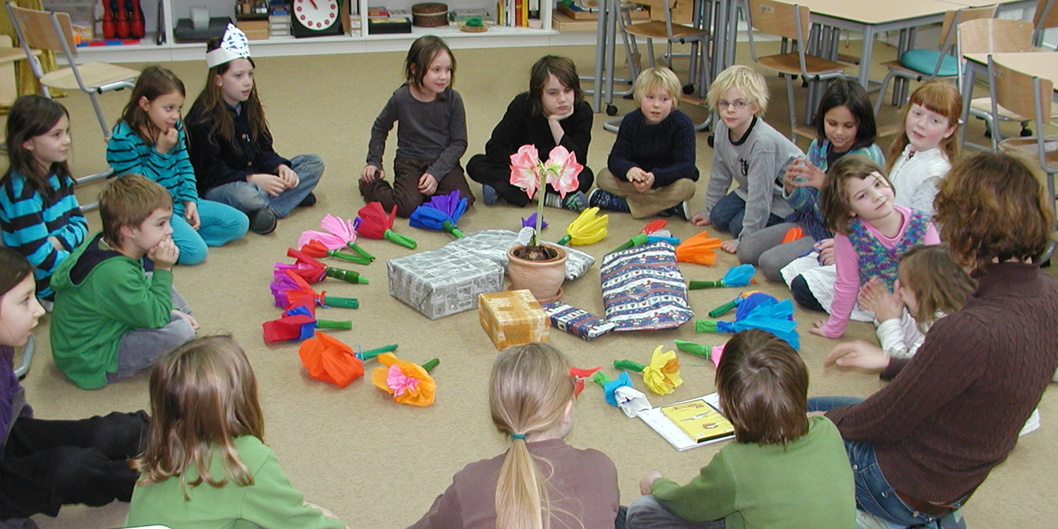 Eine Klasse sitzt in einem Kreis und in diesem Kreis befinden sich selbstgebastelte Blumen, eine echte Blume und fünf Geschenke