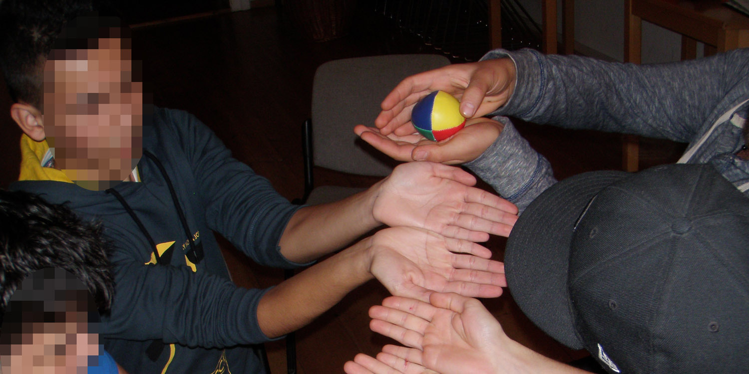 Jugendliche versuchen, gemeinsam einen Ball ihre aneinandergereihten Händer herabrollen zu lassen, ohne, dass er herunterfällt.