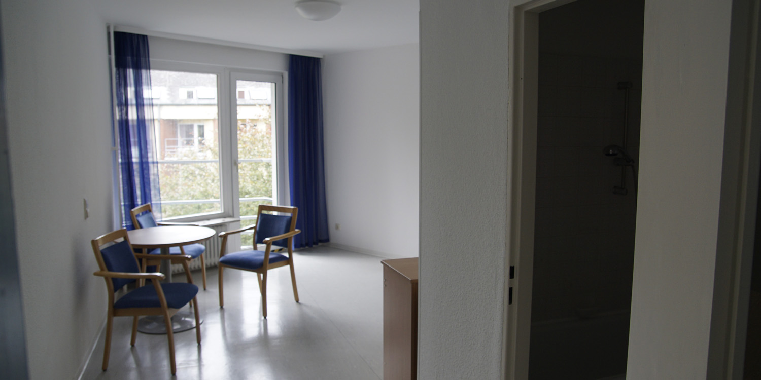 Ein Raum, der mit einem Tisch mit zwei Stühlen und einem kleinen Schrank gefühlt ist. Im Hitnergrund sieht man ein Fenster und eine Tür zum Balkon