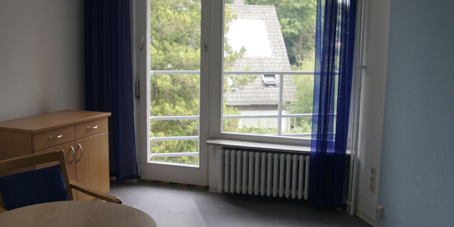 Ein Raum mit blauen Gardinen, einer Balkontür und Fenster, einen Schrank und einen Tisch mit einem Stuhl