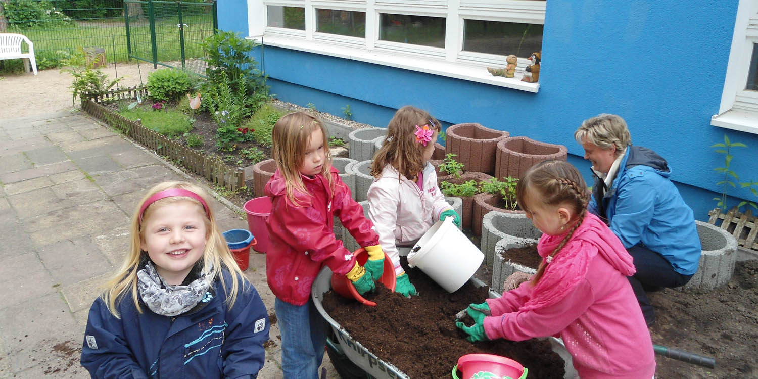 Mehrere Kinder und eine Erzieherin, die Erde aus einer Schubkarre in Eimer umfüllen. Im Hintergund sieht man mit Erde befüllt und unbefüllte Blumenkästen.