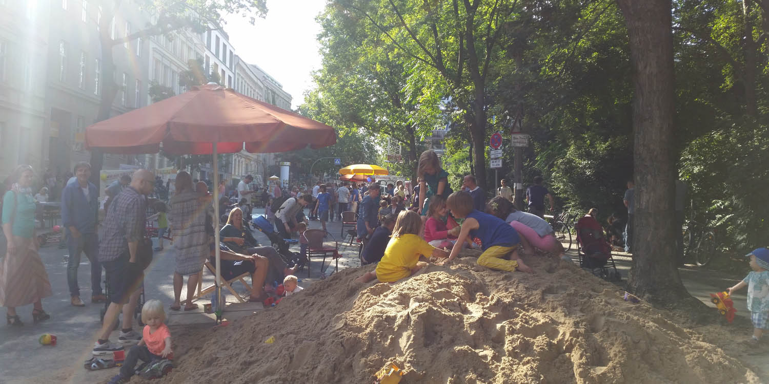 Im Hintergrund sieht man ein Fest und im Vordergrund ist ein Sandhaufen, wo Kinder spielen