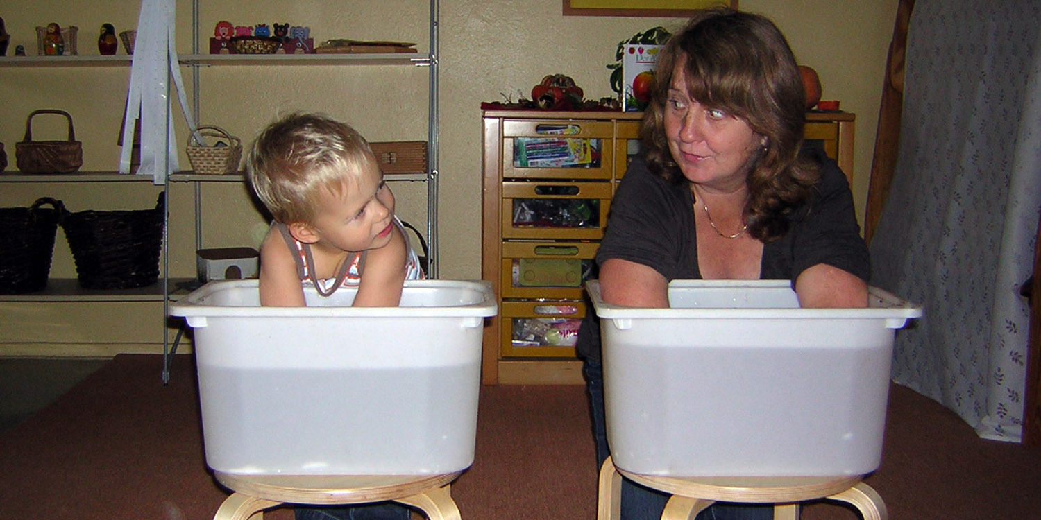 Kleiner Junge und Erzieherin halten ihre Hände in jeweils ein Becken mit Wasser.