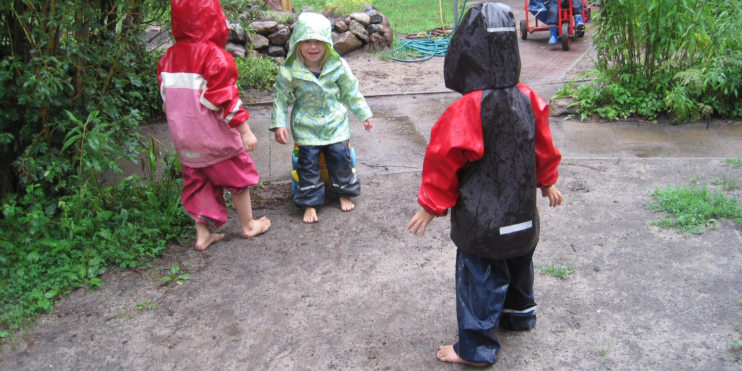 Drei kleine Kinder laufen Barfuß und in Regenkleidung auf leicht feuchter Erde