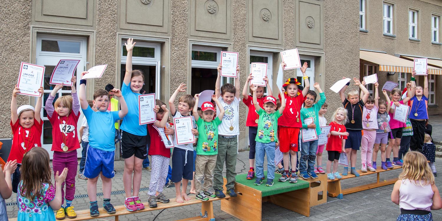 Viele Kinder, die draußen auf Sportbänken stehen und eine Urkunde hoch halten und eine Medaille um den Hals haben