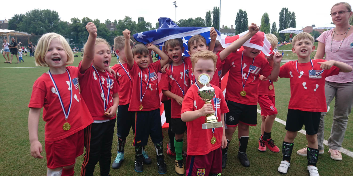 Eine Fußballmannschaft der Kita, die Haben Medaillien um den Hals und einer trägt einen Pokal. Die Kinder tragen auf ihren Trikots eine Flagge der USA und heben die Nationalflagge der USA über sich