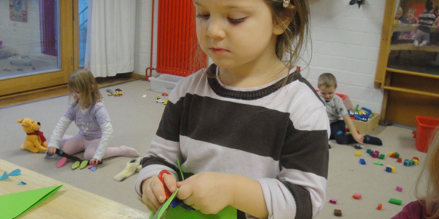 Ein Mädchen bastelt etwas und im Hintergrund sieht man zwei Kinder spielen