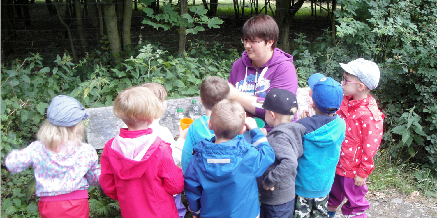 Mehrere Kinder, die vor einer Holzbank stehen und mit einer erwachsenen Person etwas trinken