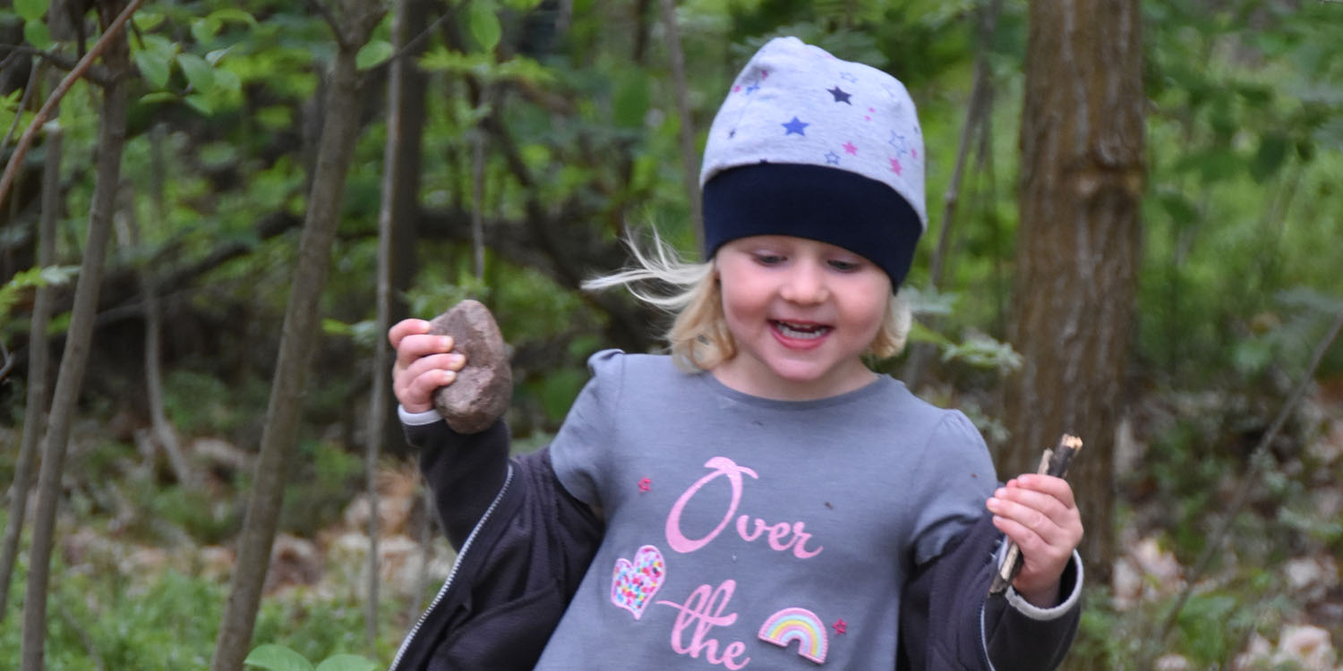 Ein Mädchen lacht und hat einen Stein und kleine Äste in der Hand im Hintergrund ist ein Wald zu sehen