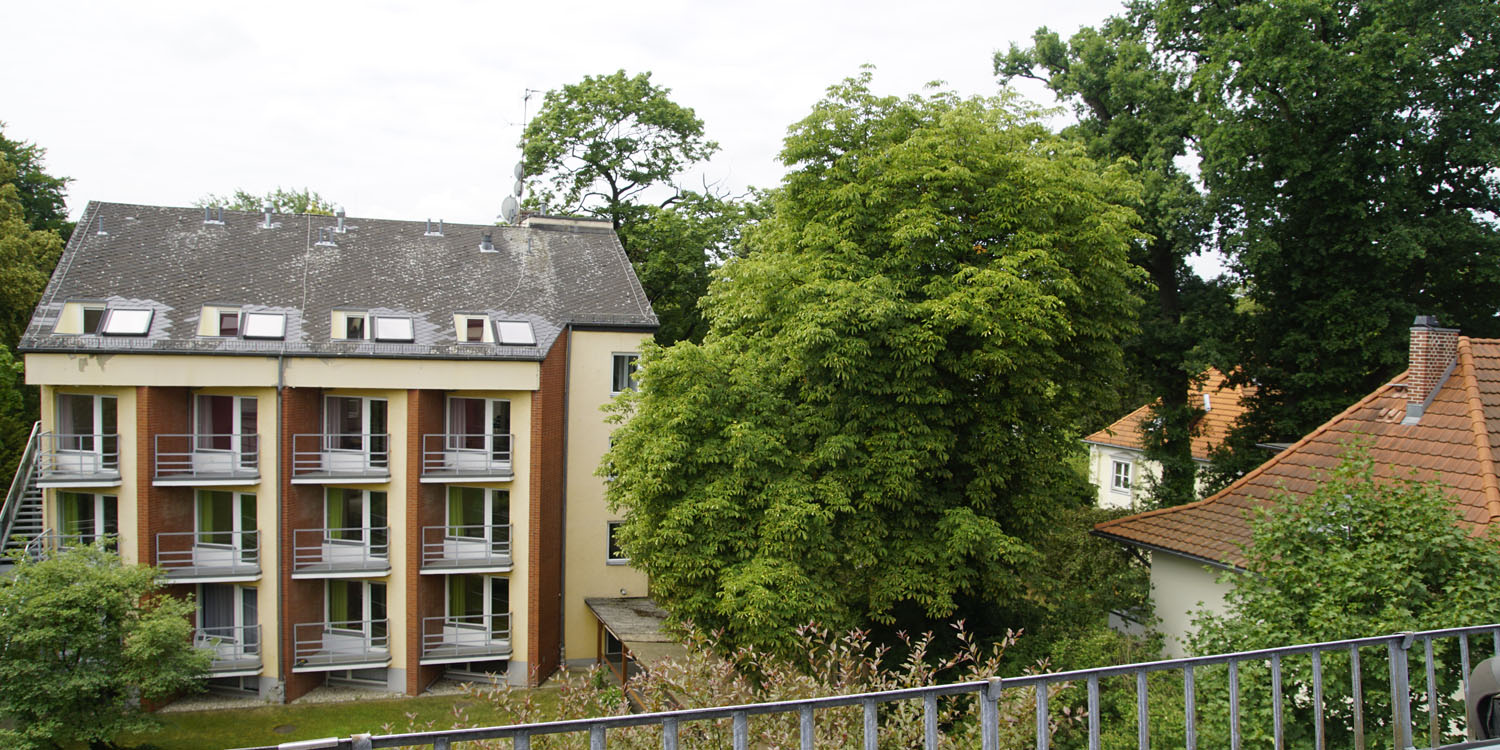 Blick aus dem Balkon auf einen grünen Garten mit Rasen und alten Bäumen sowie das Haus gegenüber