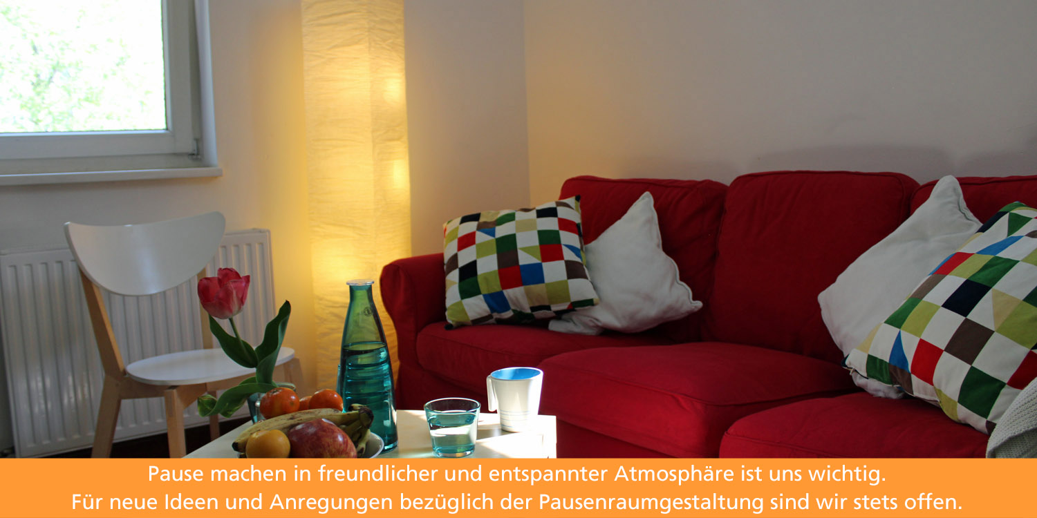 Im Pausenraum lädt ein rotes Sofa zum Entspannen ein. Auf einem viereckigen isch stehen eine Obstschale, Wasser und eine Vase mit einer Tulpe.