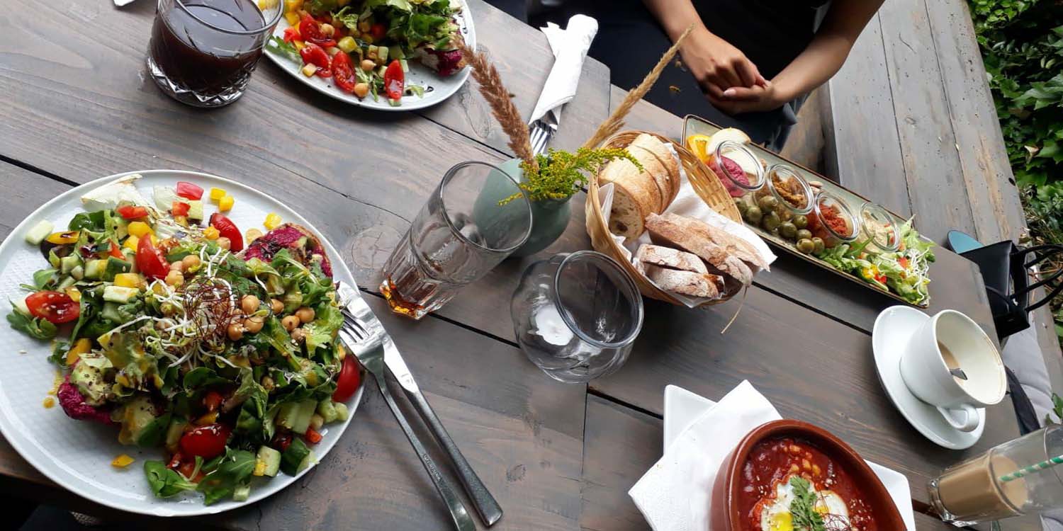 Auf einem gedeckten Holztisch stehen vier Teller mit Salat, Tomaten und Oliven. An den Tellern stehen Getränke wie Cola und Kaffee.