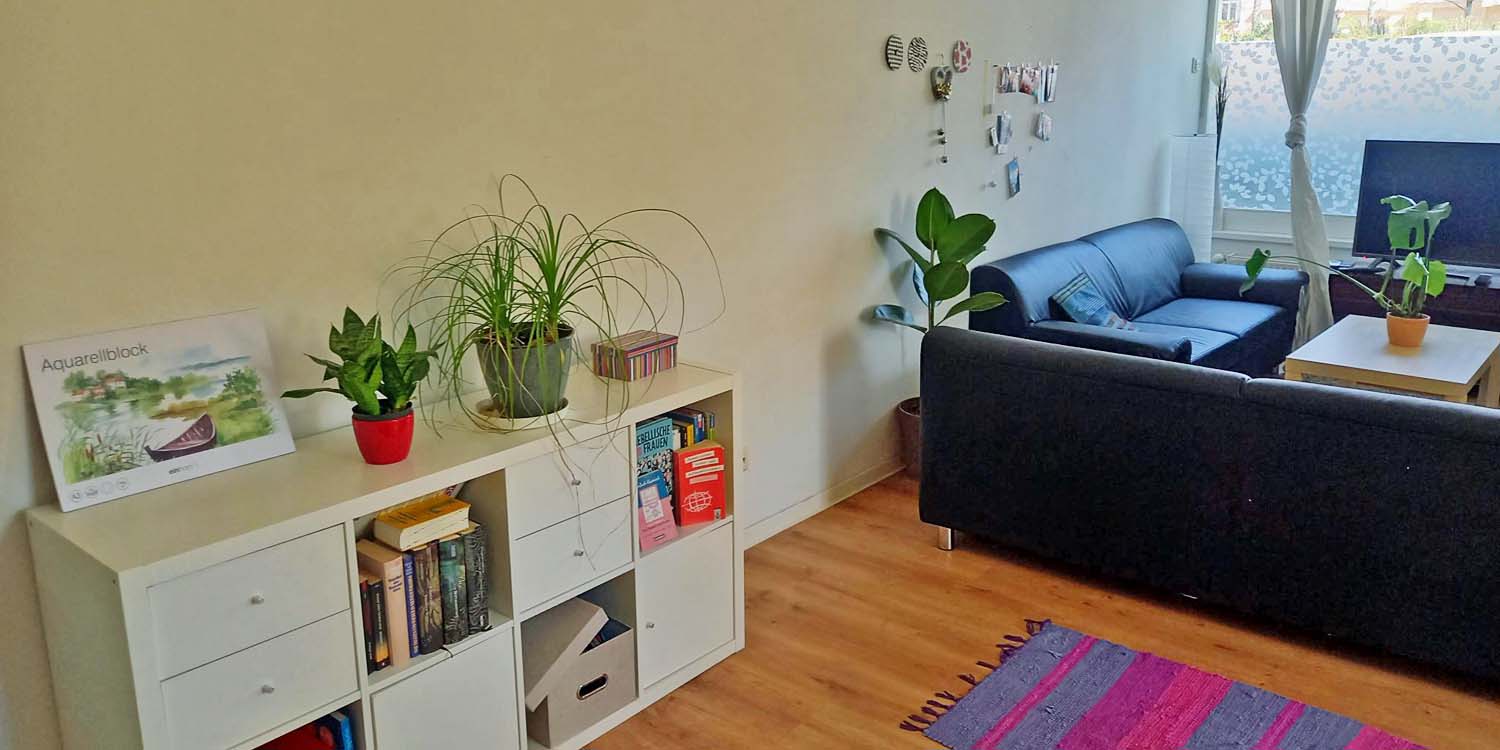 Im Vordergrund steht ein Sideboard mit Schubladen. Auf dem Seideboard sind kleine Grünpflanzen und ein Aquarellblock. Im Hintergrund steht eine Sitzecke mit Sofas und einem Holztisch ein der Mitte.