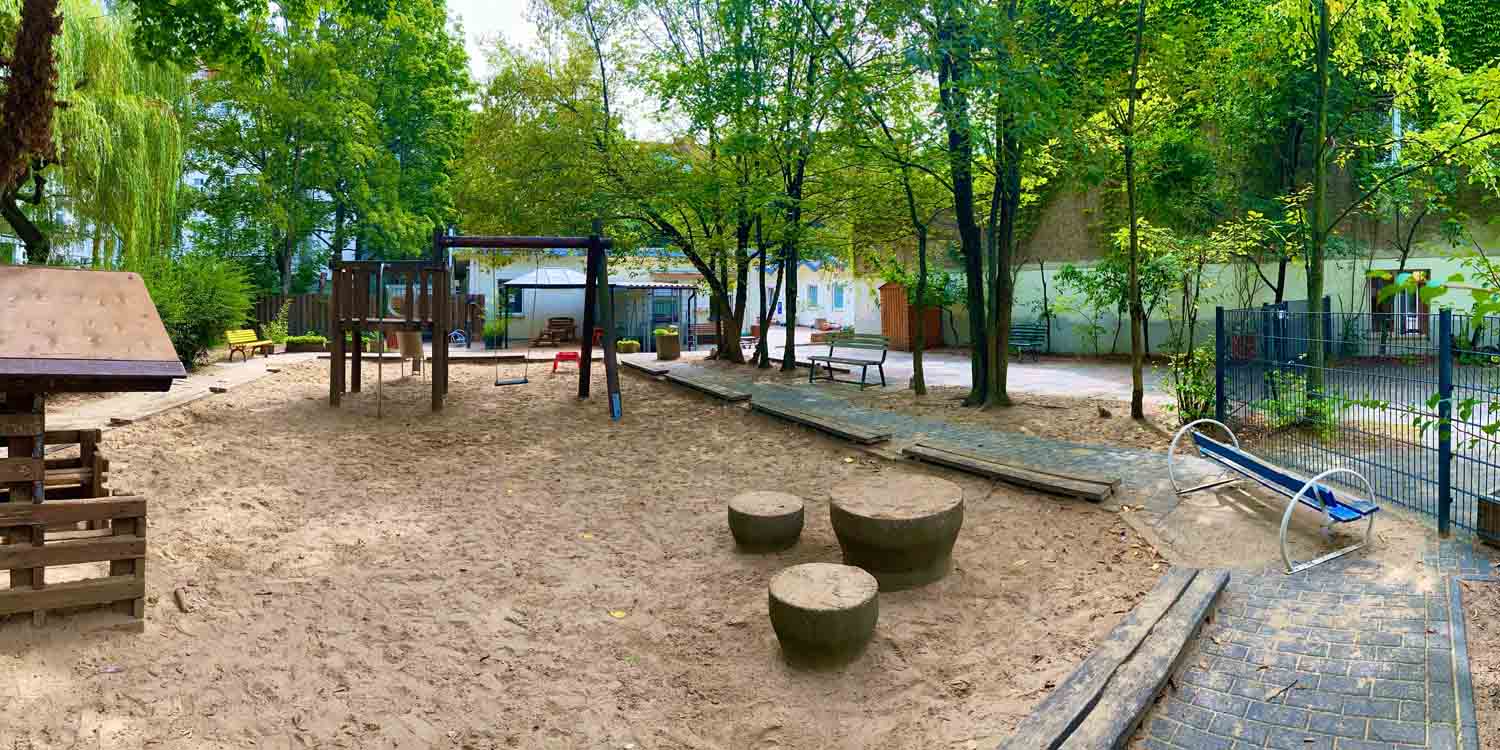 In dem großen Sandkasten stehen Schaukeln und kleine Holzstämme, die die Kinder zum Klettern anregen. Im Vordergrund steht ein kleines Holzhaus; ringsherum Bänke.