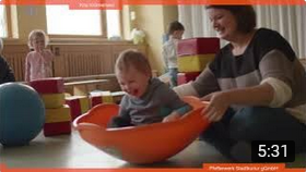 Thumbnail YouTube-Video: Ein Kleinkind sitzt in einer halbrunden Schale und wird von einer Pädagogin geschaukelt.