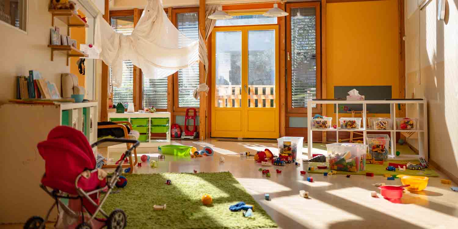 In einem sonnendurchfluteten Raum stehen mehrere weiße Regale mit Spielsachen. Im Vordergrund ein roter Puppenwagen.