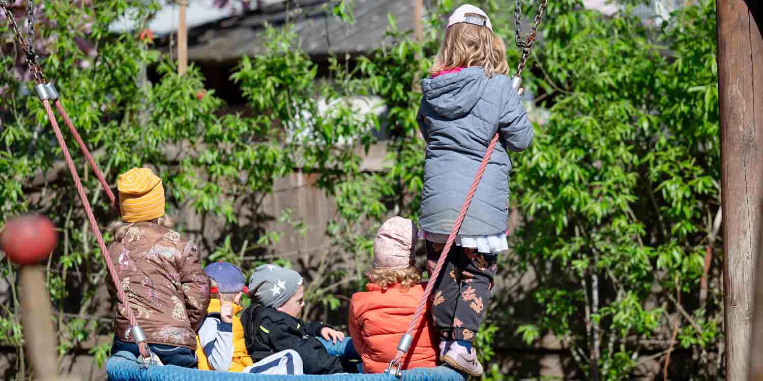 Vier Kinder sitzen in einer Nestschaukel. Ein Kind steht am Rand und hält sich am Seil fest.
