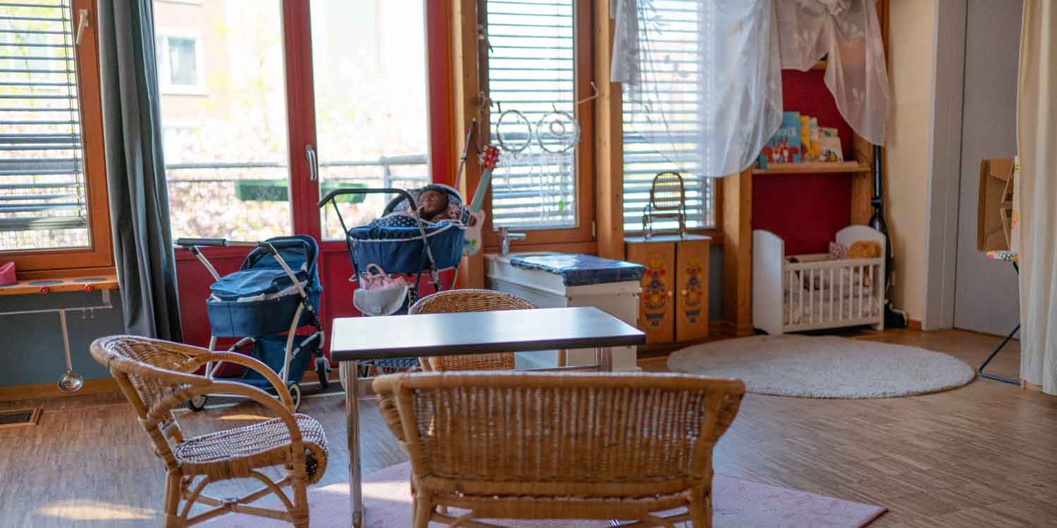 Mitten im Raum steht ein viereckiger Holztisch mit drei Korbstühlen. An der Wand stehen zwei Puppenwagen, ein kleiner Wickeltisch und ein Puppenbett.
