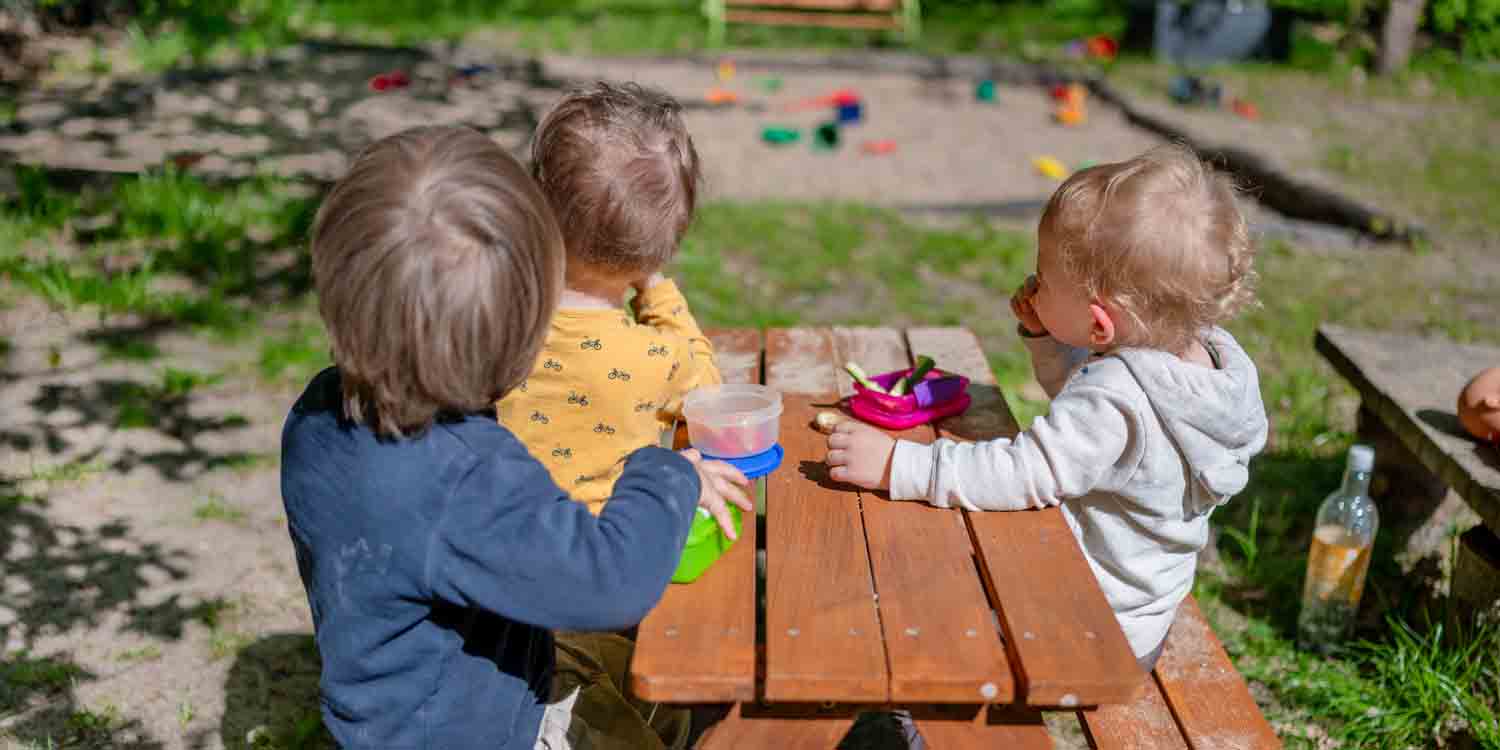 An einem kleinen Holztisch sitzen drei Kleinkinder im Garten und essen Obst.