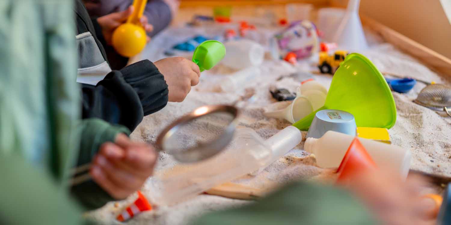 Kinderhände halten Trichter und kleine Schaufeln und spielen im Sand.