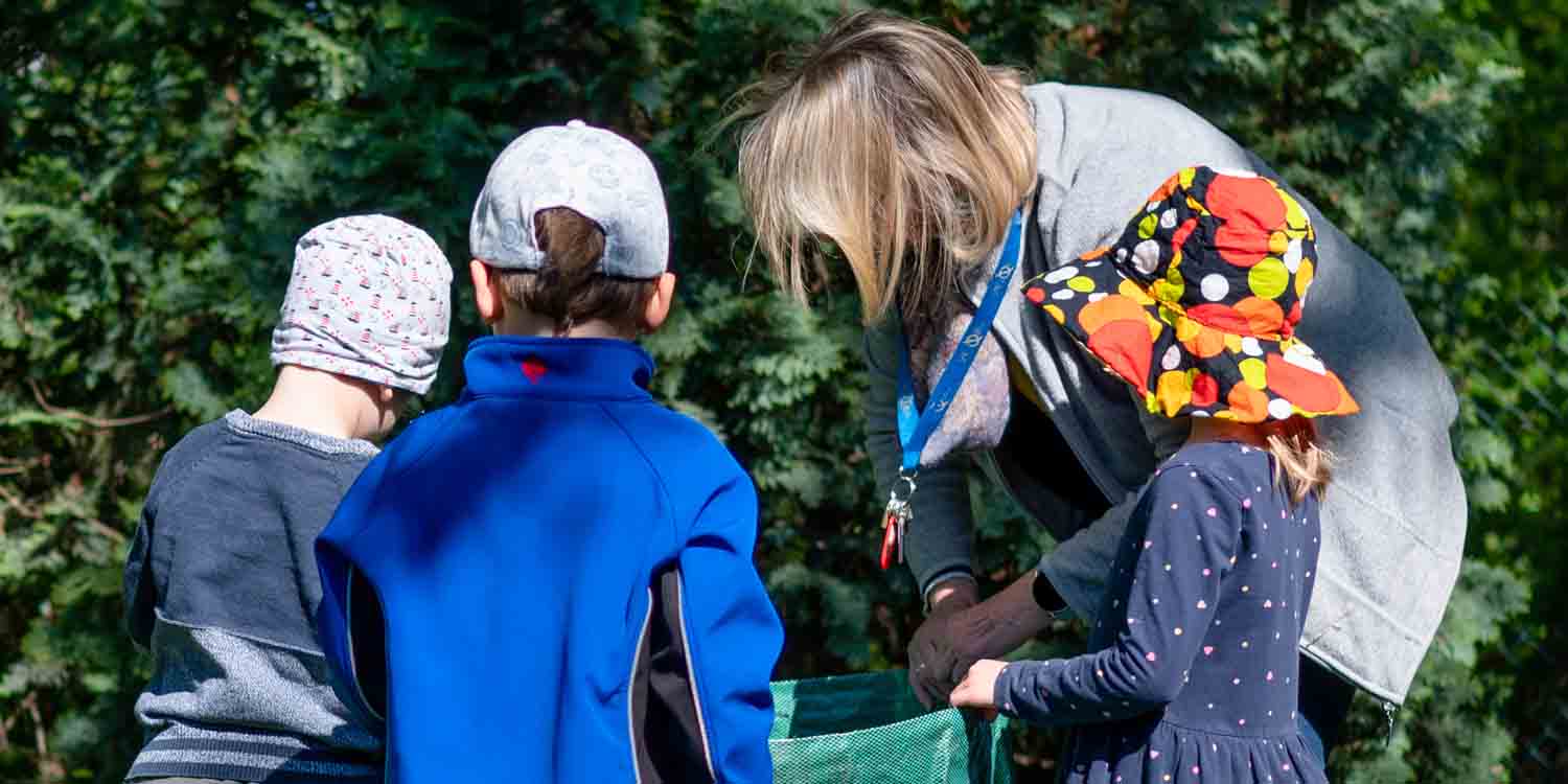 Drei Kita-Kinder und eine pädagogische Fachkraft sammeln Müll in einem Park und werfen ihn in eine große grüne Tüte.