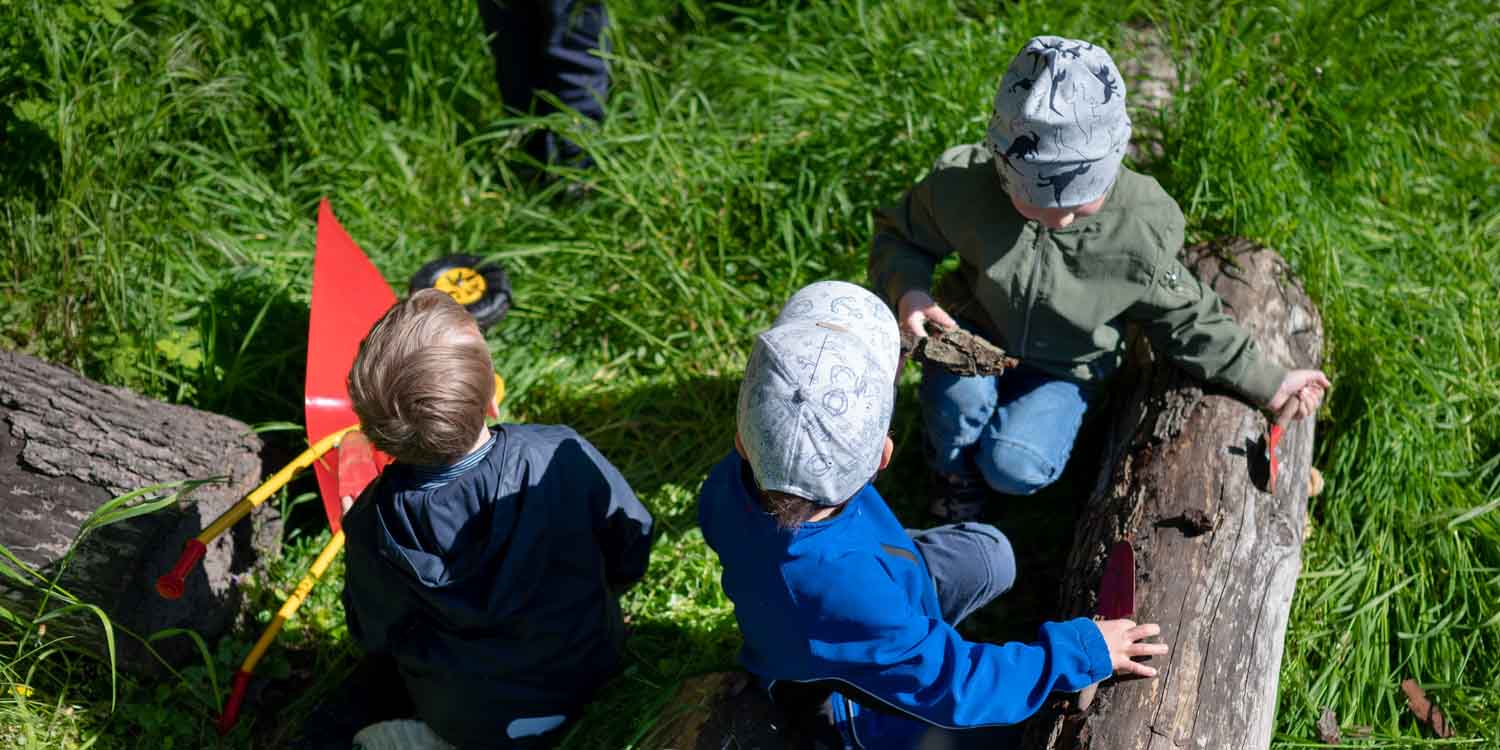 Drei Kita-Kinder sitzen im Gras und legen im Garten Gefundenes auf zwei auf dem Boden liegende Baumstämme.