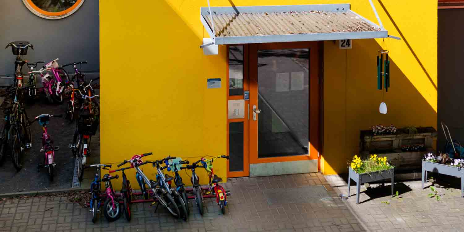 Vor dem gelb gestrichenenen Eingang stehen viele Kinderfahrräder und Roller. Rechts neben der Eingangstür stehen zwei Blumenkästen mit blühenden Blumen, über denen ein Klangspiel aus Metall hängt.