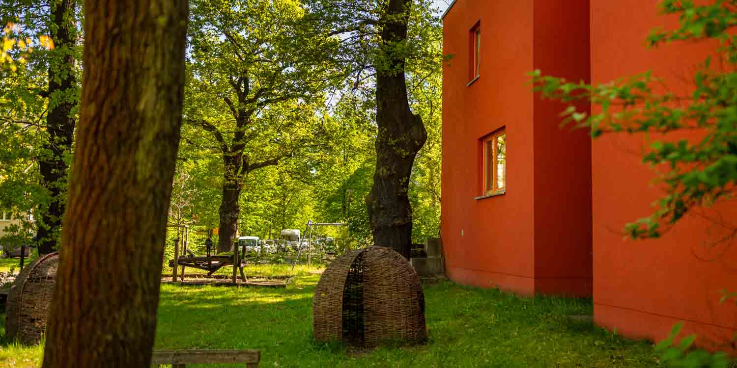 Im Kita-Garten steht vor dem roten Anbau eine kleine runde Hütte aus Korbgeflecht. Im Vordergrund ragt ein Baumstamm ins Bild.