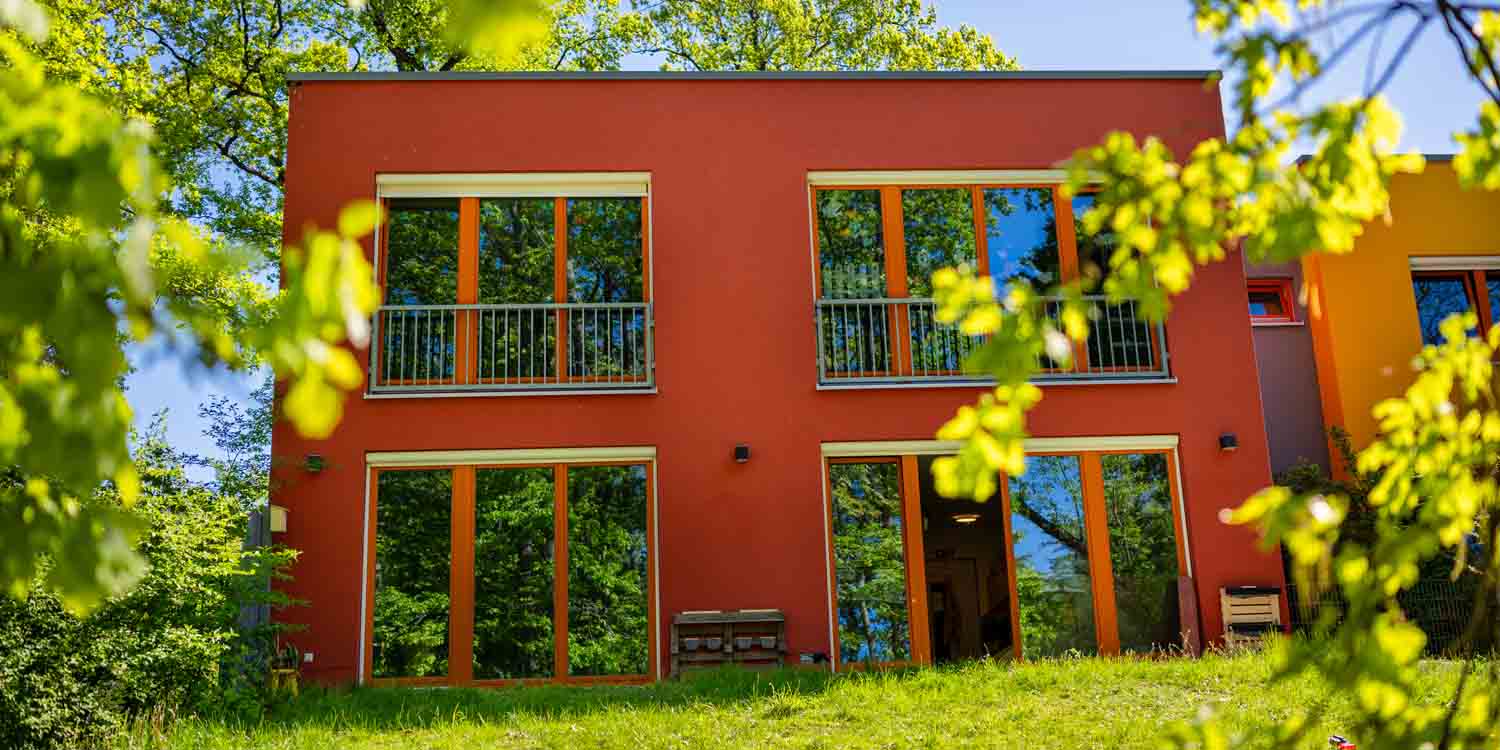 Der ziegelrote Kita-Anbau steht auf der Wiese im Kita-Garten und wird umrahmt von ins Bild hineinrankenden Blättern.