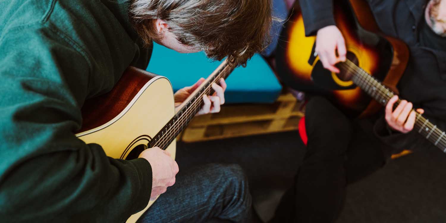 Zwie jungen Menschen spielen Gitarre und beugen sich dabei über die Instrmente, um die Saiten zu greifen.