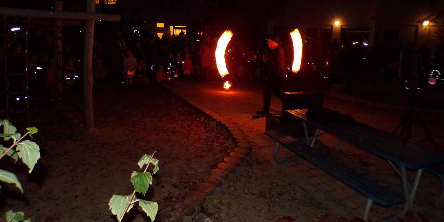 Im Kita-Garten steht im Kreis der Eltern und Kinder ein Mensch, der mit Fackeln ein Feuershow vorführt. Es ist dunklen und die Fackeln bilden durch das Herumschleudern einen Halbkreis.