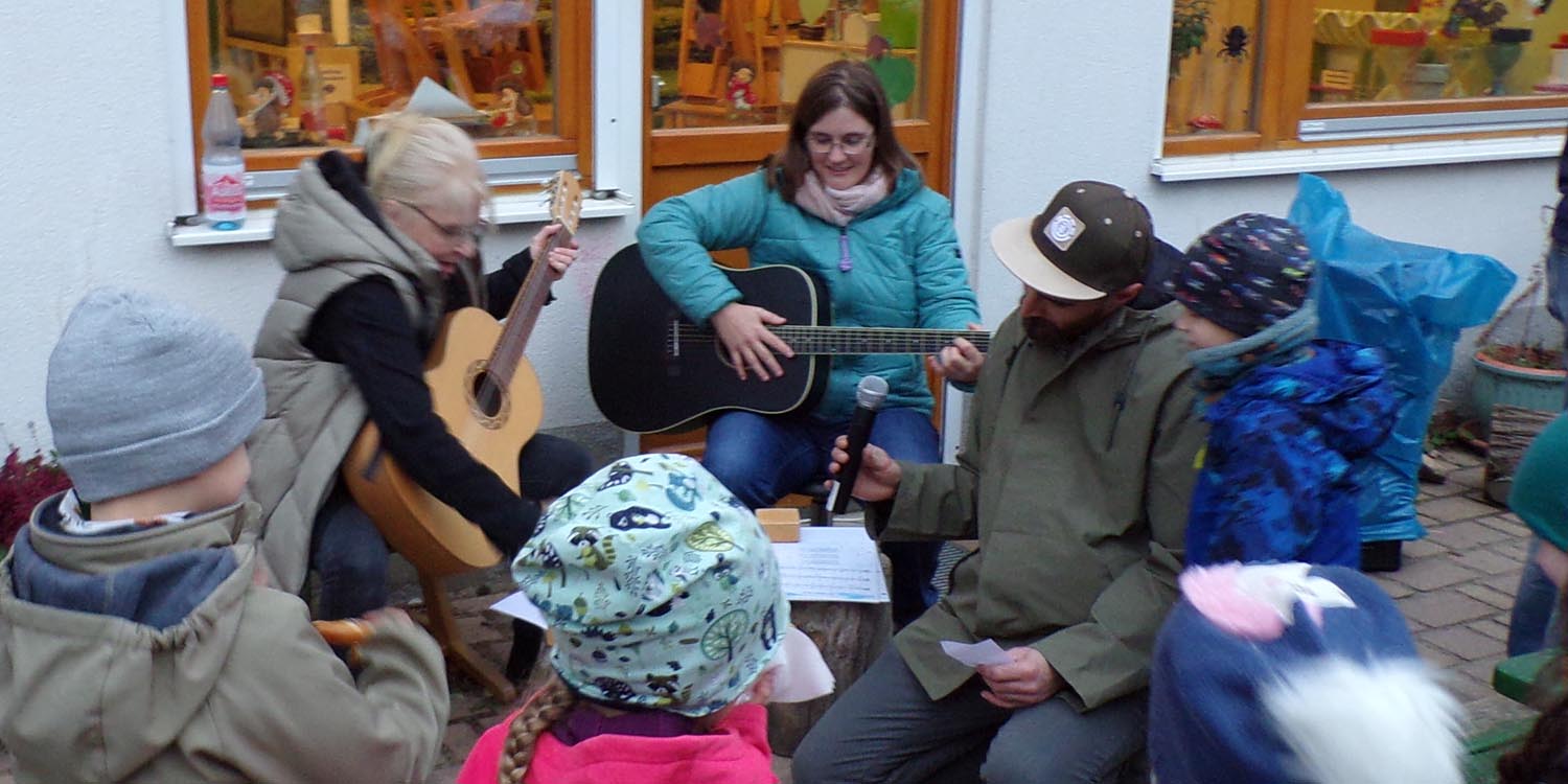 Zwei pägagogische Fachkräfte spielen Gitarre; Kinder stehen im Kreis herum.