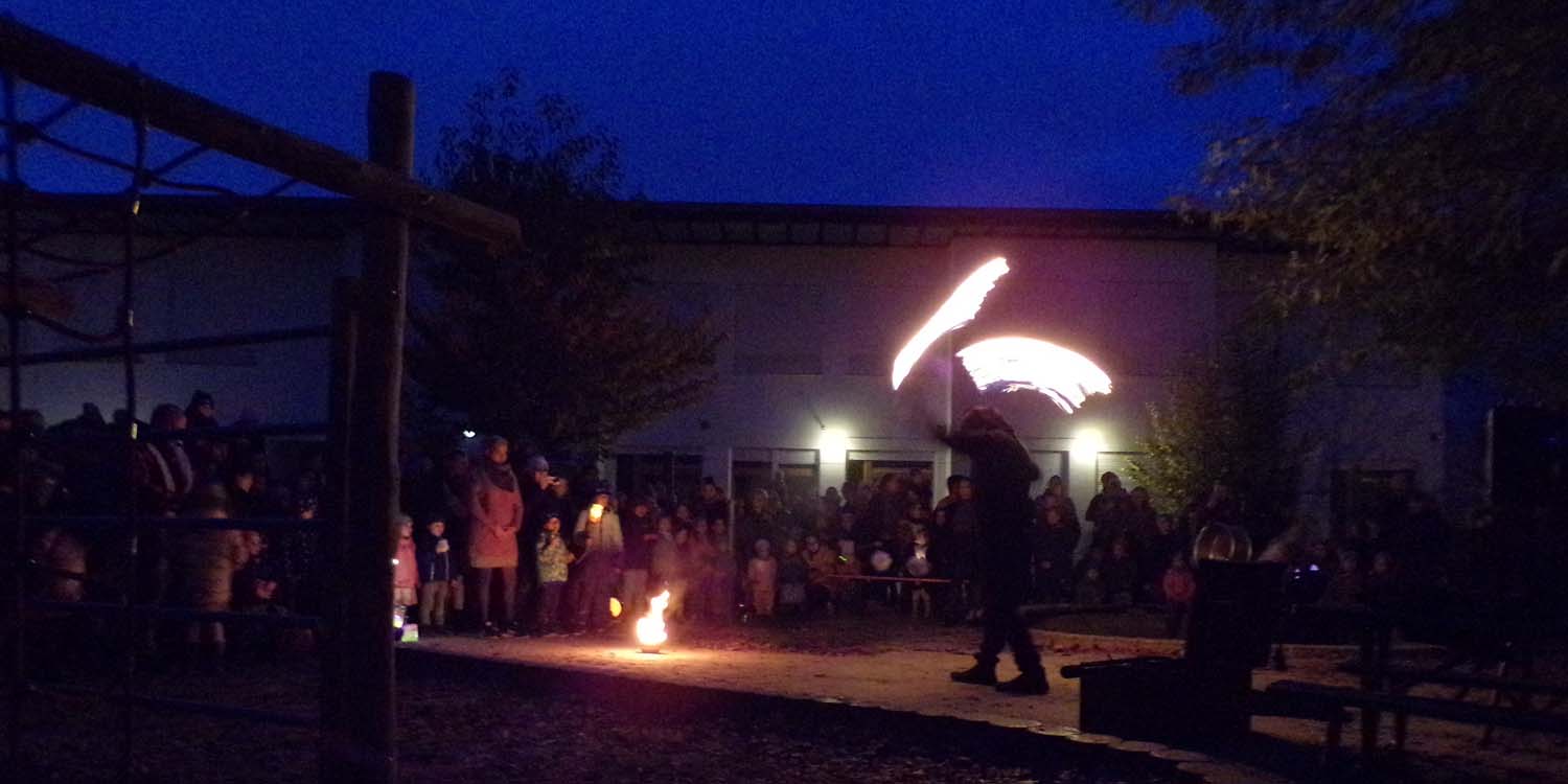 Im Kita-Garten steht im Kreis der Eltern und Kinder ein Mensch, der mit Fackeln ein Feuershow vorführt. Es ist dunklen und die Fackeln bilden durch das Herumschleudern einen Halbkreis.