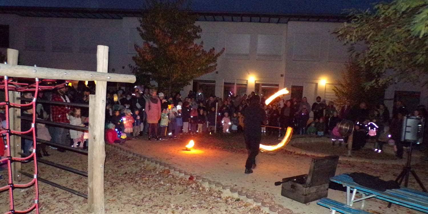 Im Kita-Garten steht im Kreis der Eltern und Kinder ein Mensch, der mit Fackeln ein Feuershow vorführt. Es ist dunklen und die Fackeln bilden durch das Herumschleudern einen Halbkreis.