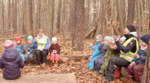 Kinder und Pädagog*innen sitzen auf zwei umgestürzten Bäumen im bunt gefärbten Herbstwald und winken sich zu.