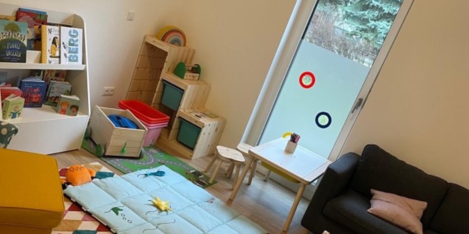 In einem Raum stehen ein Bücherregal mit Kinderbüchern, ein kleiner Tisch mit zwei Kinderstühlen und in der Mitte eine MAtte mit Spielsachen. Im Hintergrund ein Fenster, das bis zum Boden reicht und in der Ecke ein weiteres Regal.