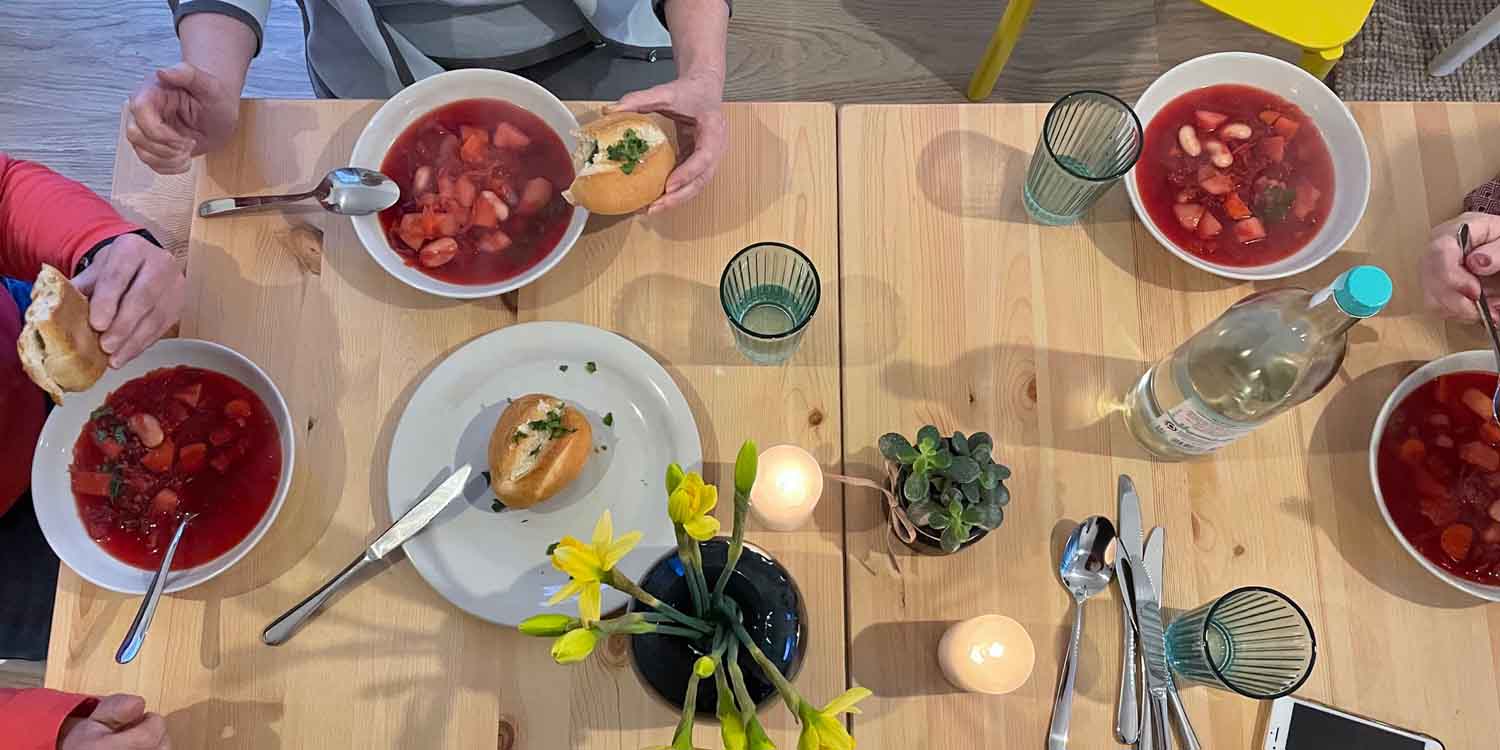 Ein Tisch von oben mit vier Schüsseln Borschtsch und einem Teller mit Brötchen. Dazwischen stehen Gläser, eine Wsserflasche und eine Vase mit Osterglocken.