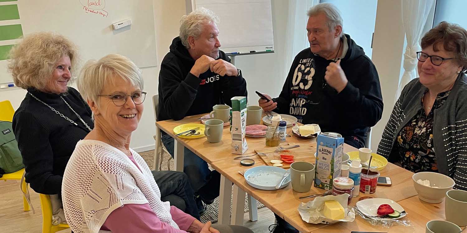 Fünf ältere Menschen sitzen an einem Tisch und essen gemeinsam. Auf dem Tisch stehen Miclh, Kaffee, Marmelade und Butter.