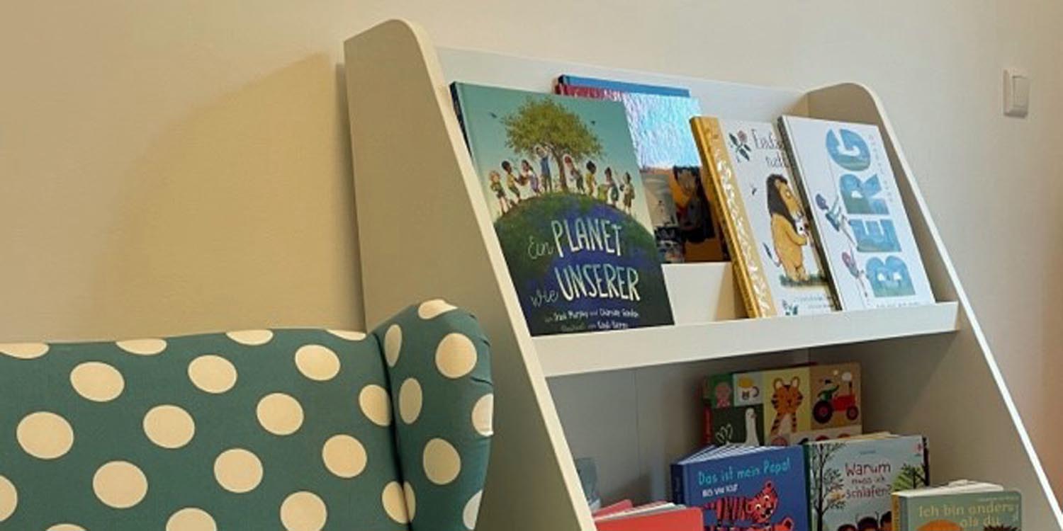 In einem Bücherregal stehen Kinderbücher in zwei Reihen. Links daneben ein gepunkteter Lesesessel.