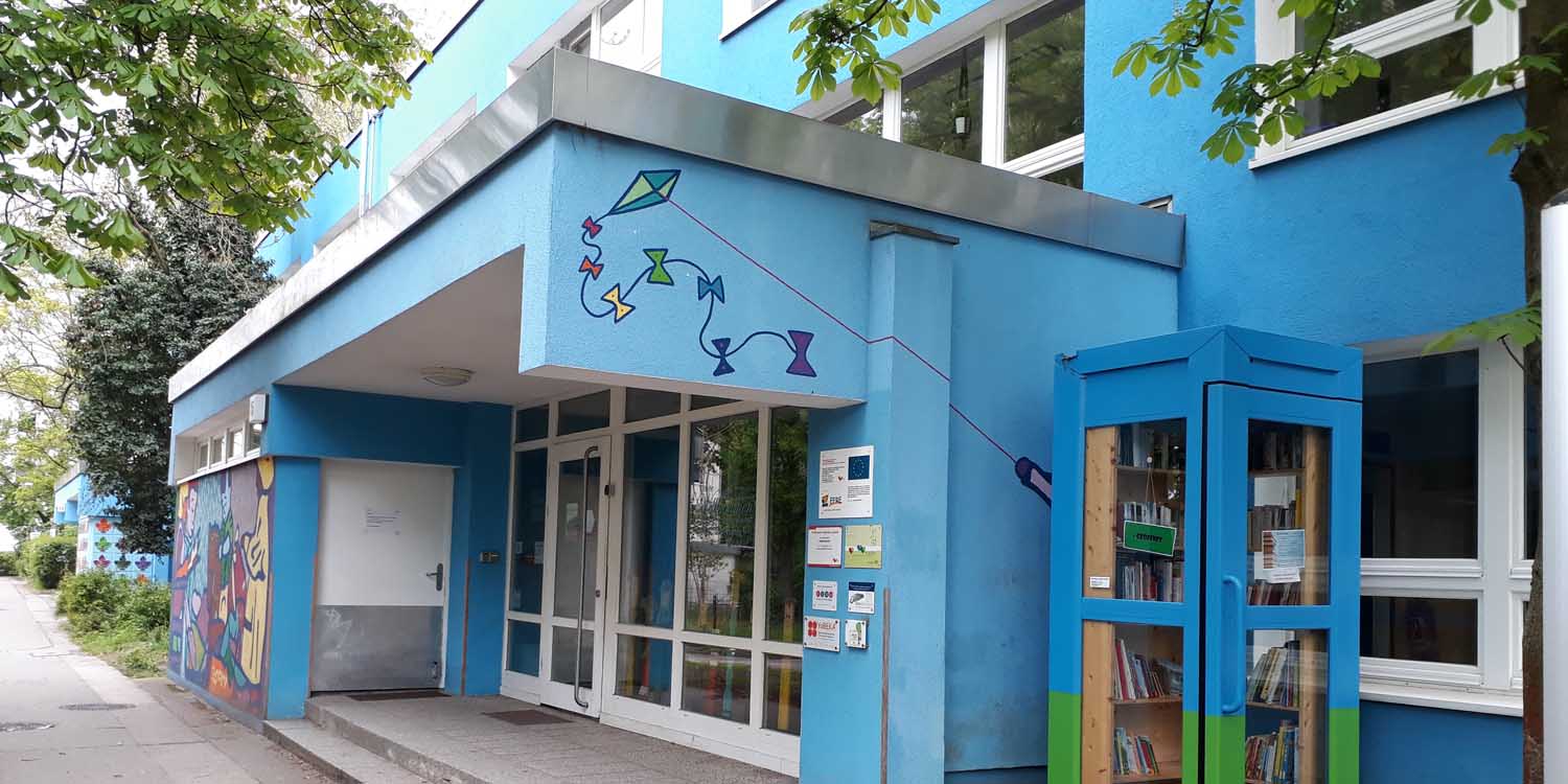 Blau gestrichener Eingang der Kita mit großen Fenstern und zwei Stufen. Rehcts neben dem Eingang steht eine ebenfalls blau angemalte Telefonzelle, in der Bücher und Spiele zum Mitnehmen und Tauschen bereitliegen. 