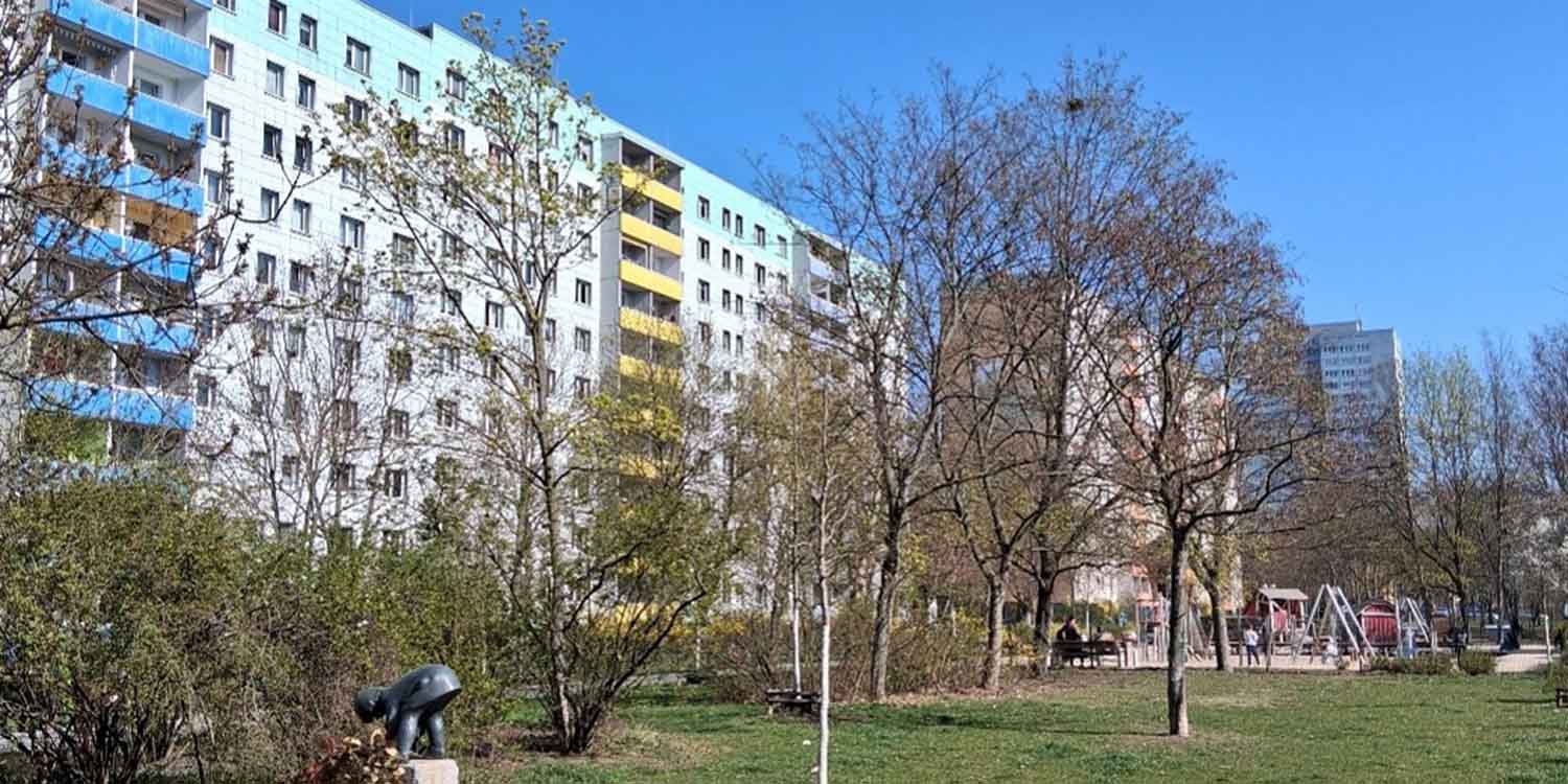 Um einen Spielplatz mit einer Grünfläche herum sind Hochhäuser gruppiert. Auf der Grunfläche stehen Bäume mit frühlingshaften Ansätzen von Blättern.