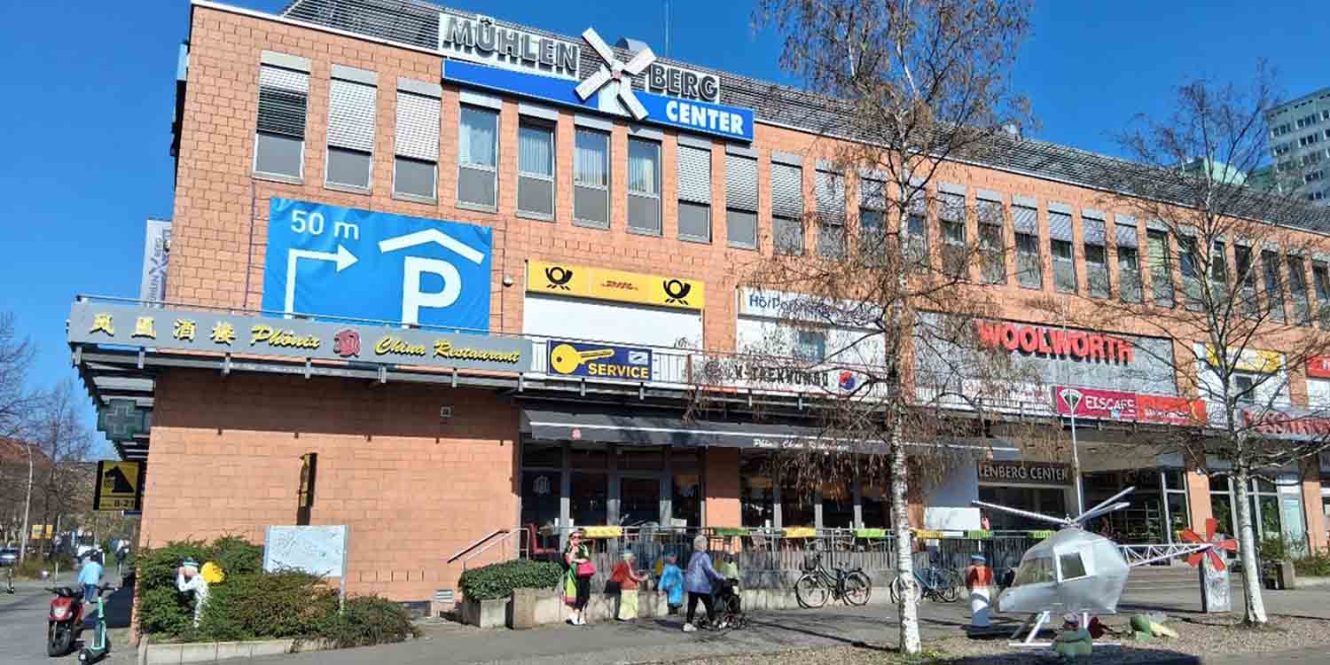 Das Mühlenbergcenter im Mühlenkiez wird von seiner Vorderansicht gezeigt. Aufschriften wie Phönix, DHL, Woolworth und Rossmann verraten, welche Geschäfte in dem Center sind. Vor dem Center sind einige Passanten unterwegs.
