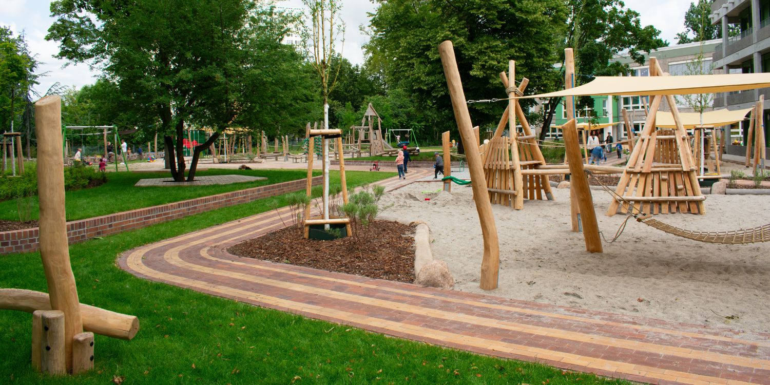 Klettergerüste in Tipiform stehen zum Teil unter Sonnensegeln im Sandkasten. Kinder laufen auf den Wegen.