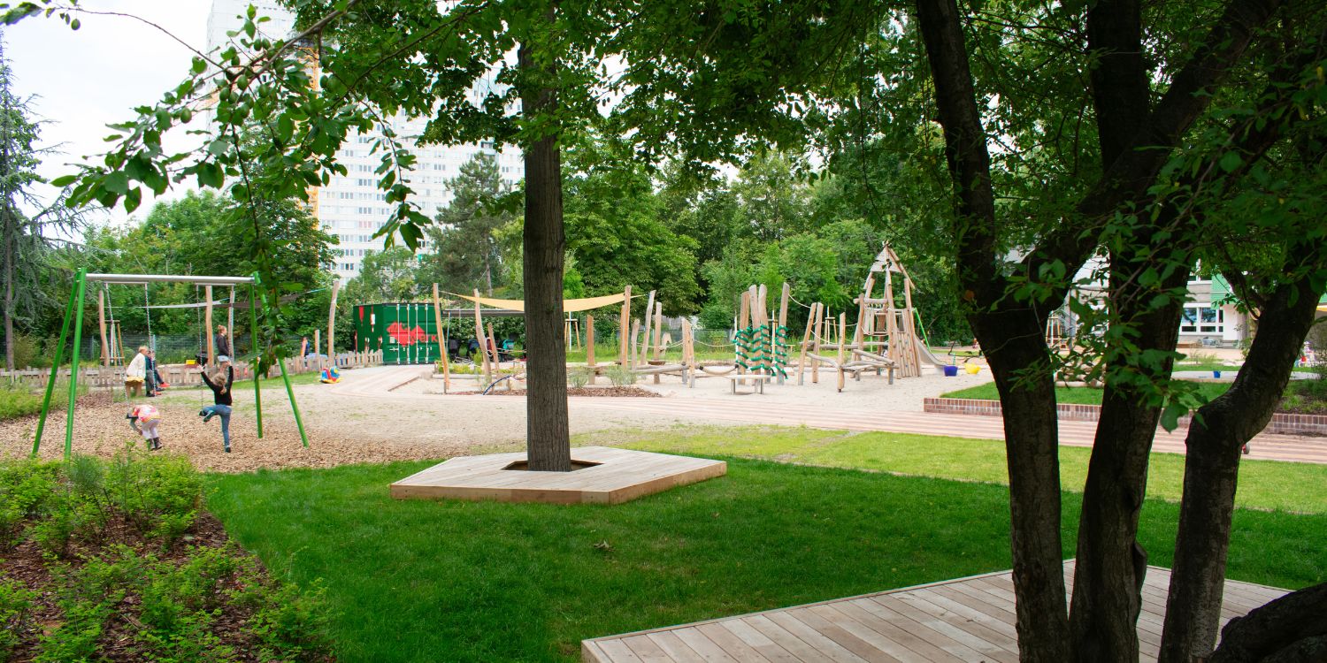 Auf einer grünen Wiese stehen große Bäume, die mit Baumscheiben umrandet sind, auf denen die Kinder sitzen können. Im Hintergrund Klettergerüste und eine Schaukel. auf der Kinder toben.