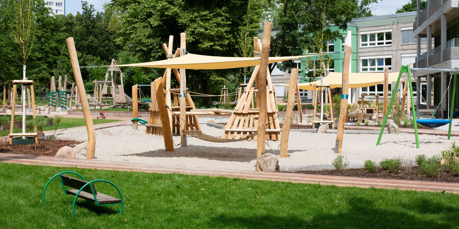 Holzgeräte und Holzklettergerüste stehen unter großen Sonnensegeln im Spielsand. Im Hintergrund steht das Kitagebäude.