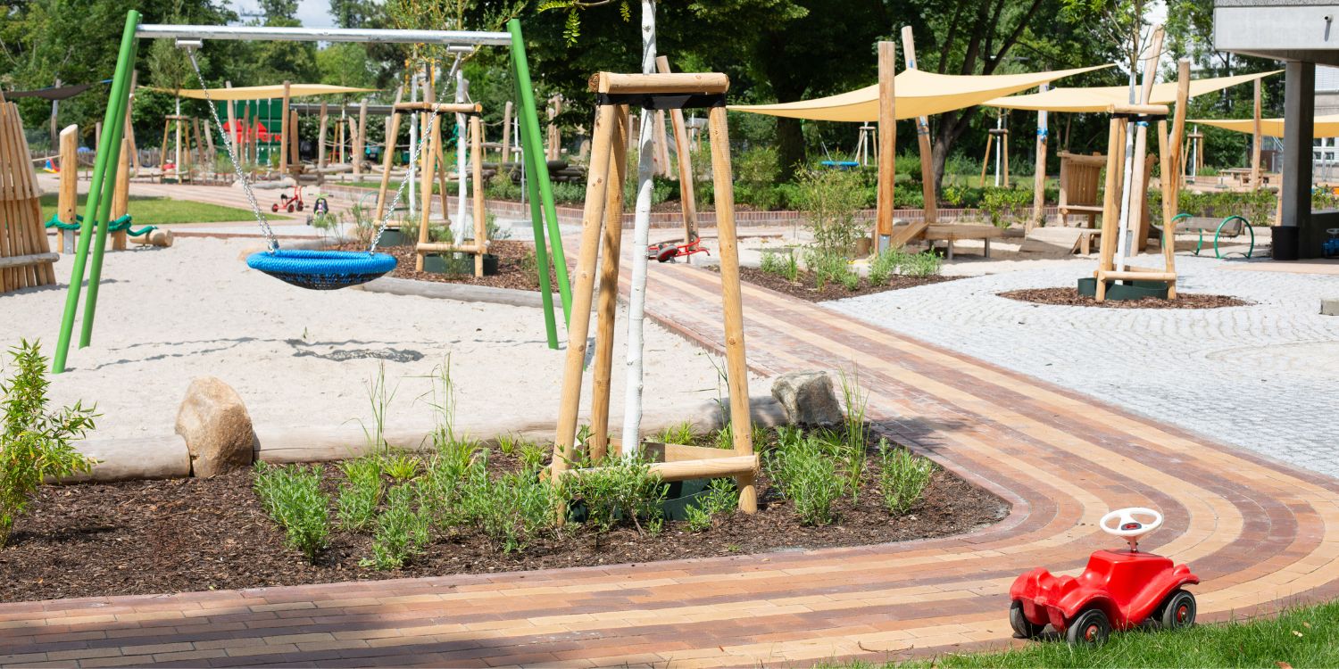 Holzgeräte und Holzklettergerüste stehen unter großen Sonnensegeln im Spielsand. Im Hintergrund steht das Kitagebäude.